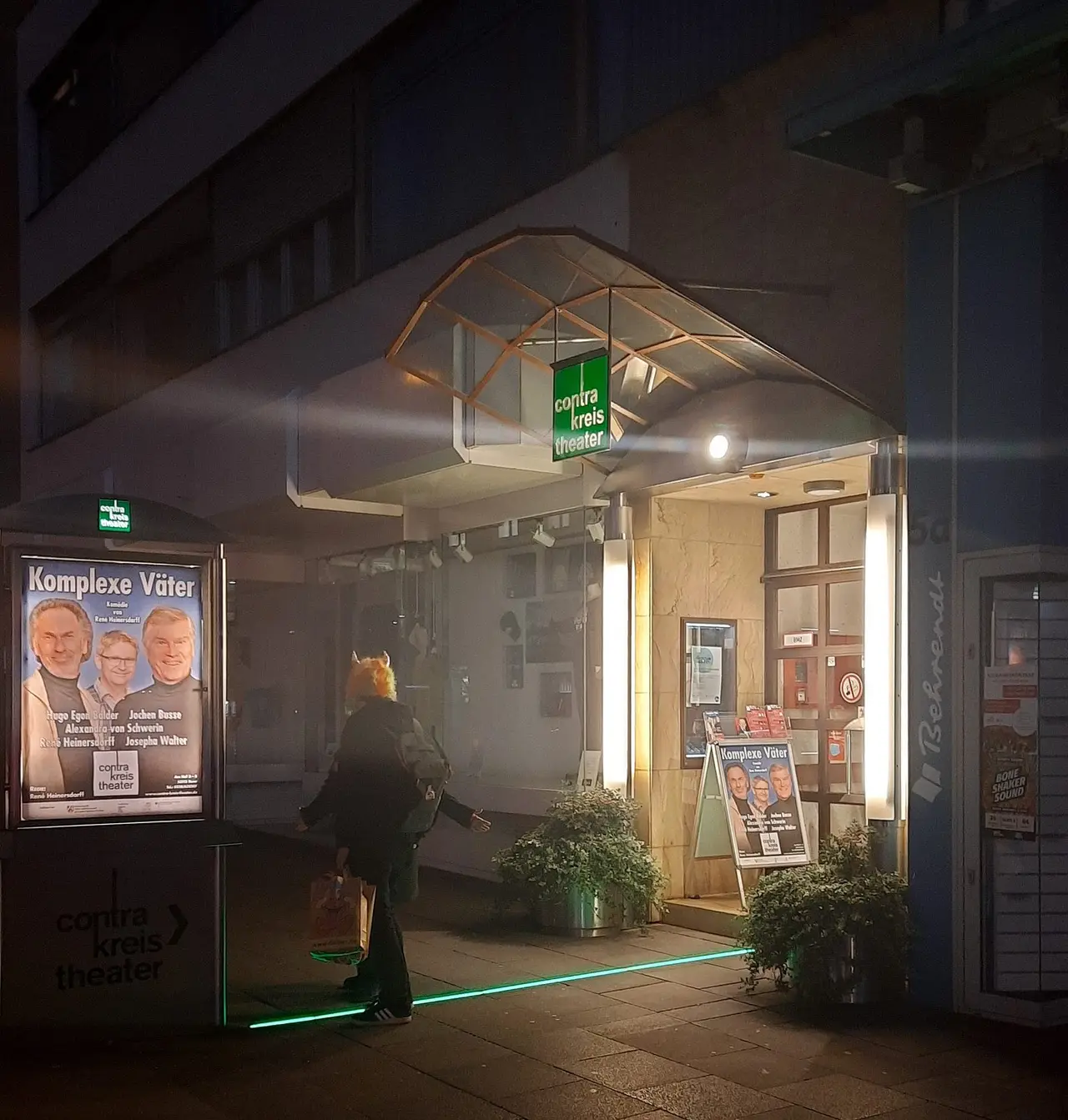 Beleuchteter Eingang des Contra-Kreis-Theaters mit Glasvordach und grünem Theaterschild