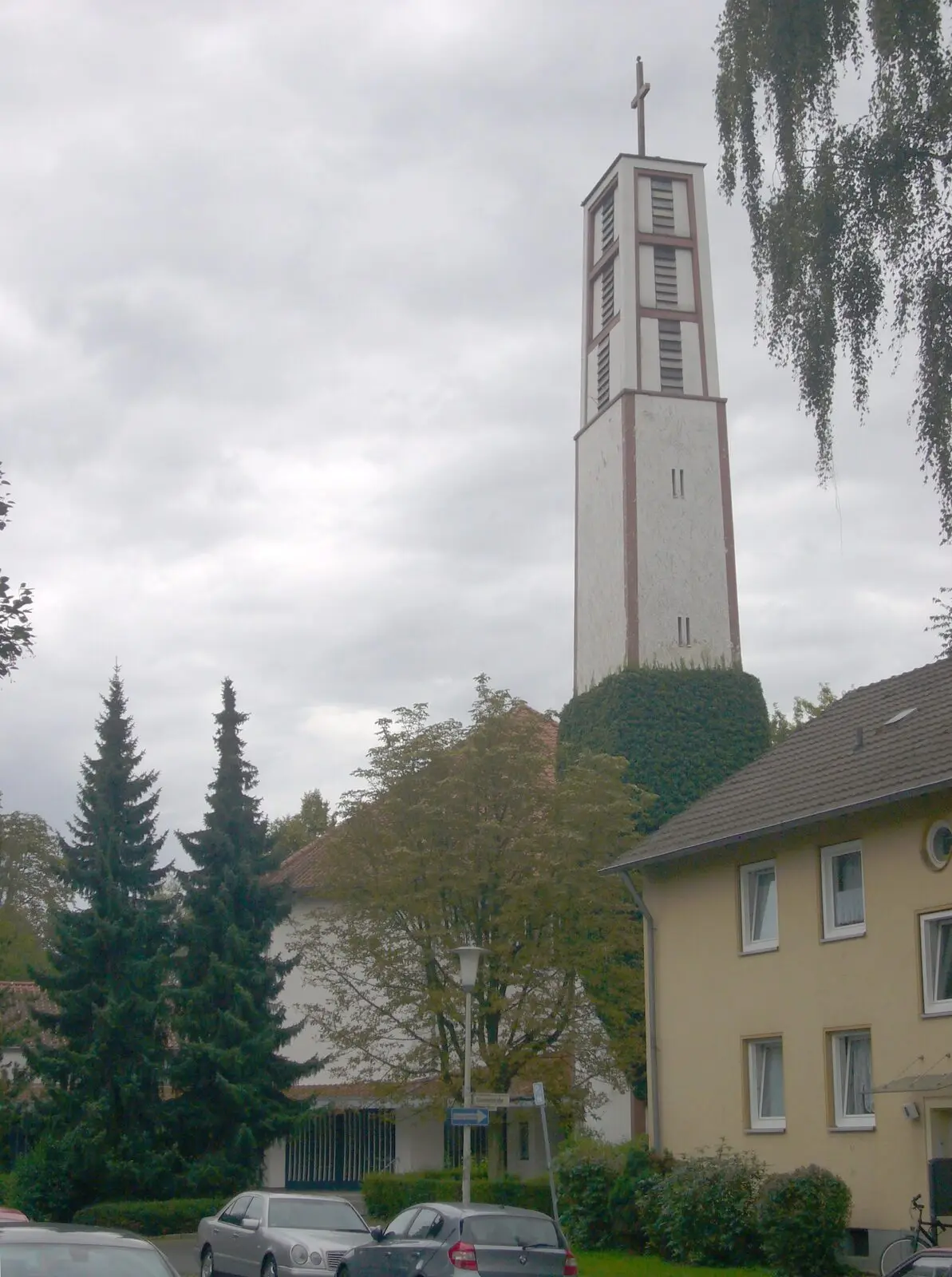Schlanker Glockenturm der Friedenskirche zwischen Nadelbäumen in Kessenich