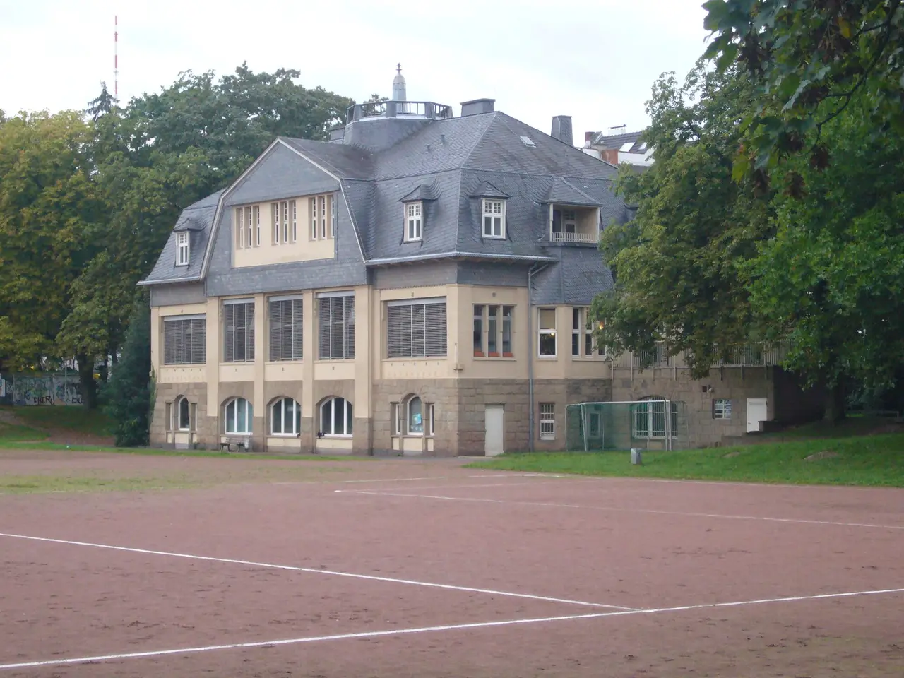 Zweigeschossiges Gebäude mit Mansarddach des Hauses der Jugend am Reuterpark, davor ein Hartplatz