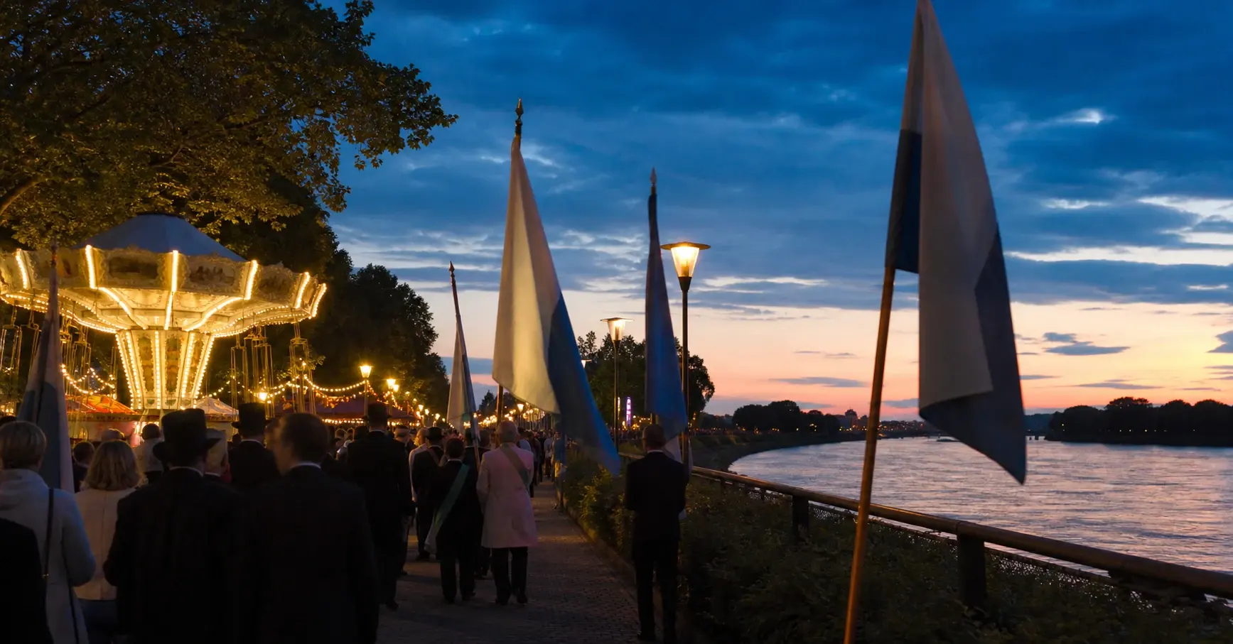 Traditioneller Festzug mit Fahnen neben beleuchteten Kirmesgeschäften am Rhein bei Abendlicht