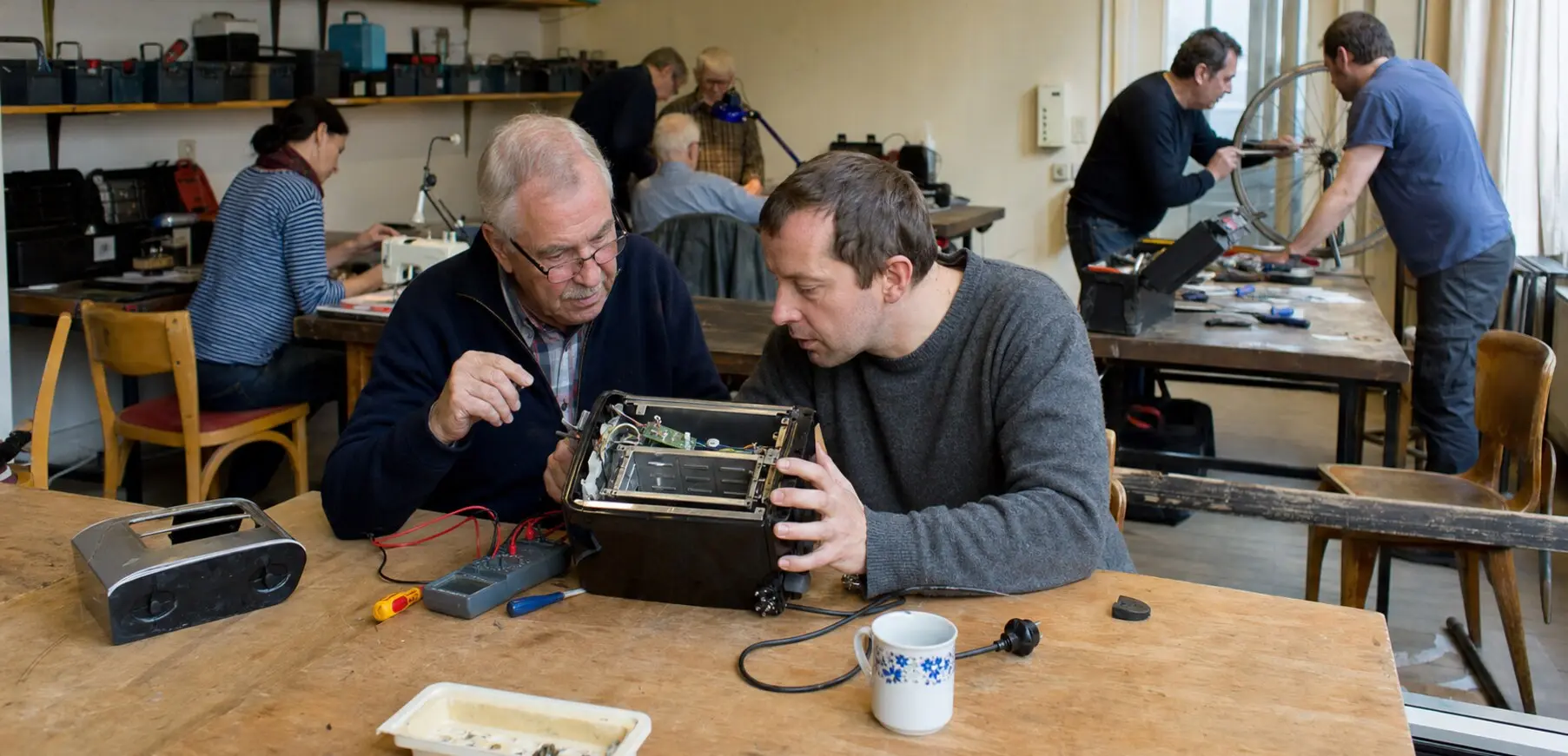 Zwei Besucher untersuchen gemeinsam ein geöffnetes Elektrogerät in einer ehrenamtlichen Reparaturwerkstatt